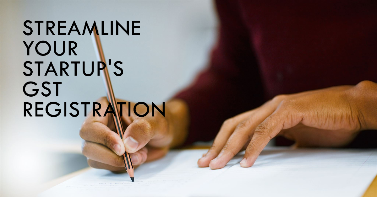 A person sitting at a desk writing in a notebook with a pencil. Text on the desk reads 'STREAMLINE YOUR STARTUP'S GST REGISTRATION.